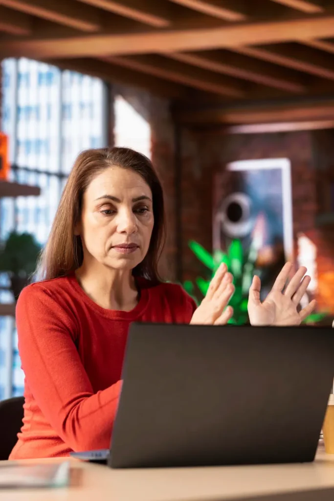 A woman in a red sweater gestures with her hands during a video call, sitting at a table with a laptop. The room has large windows and modern decor.